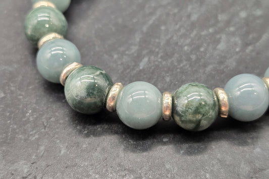 A close-up of a beaded strand featuring polished light blue-green stone beads and dark green marbled beads separated by small metallic spacer rings, set against a dark gray textured background. A watermark for 'luki lapidary' is visible in the lower-left corner.