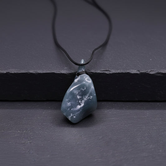 A hand holding a raw, polished gray stone pendant with a black cord, set against a blurred natural background with green moss and a gray rock.