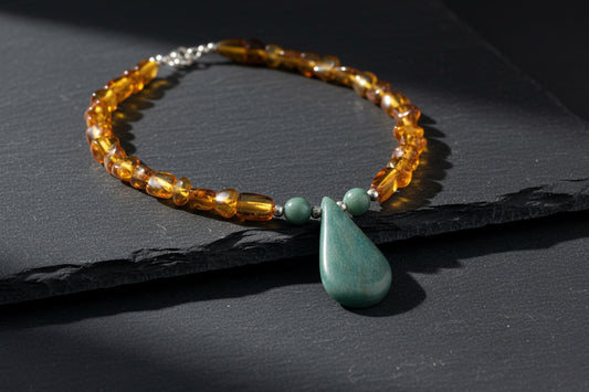 A handmade beaded necklace with a large teal-green teardrop pendant, amber-colored translucent beads, and small green spacer beads, displayed on a weathered piece of wood.