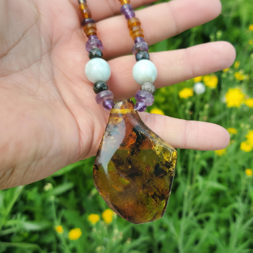 A hand holds a raw, polished amber stone pendant and jade and amethyst necklace with faceted beads, set against a blurred green grassy field with yellow flowers. A