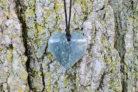 A heart-shaped light blue stone pendant with white speckles, hanging from a black cord against a textured tree bark background.