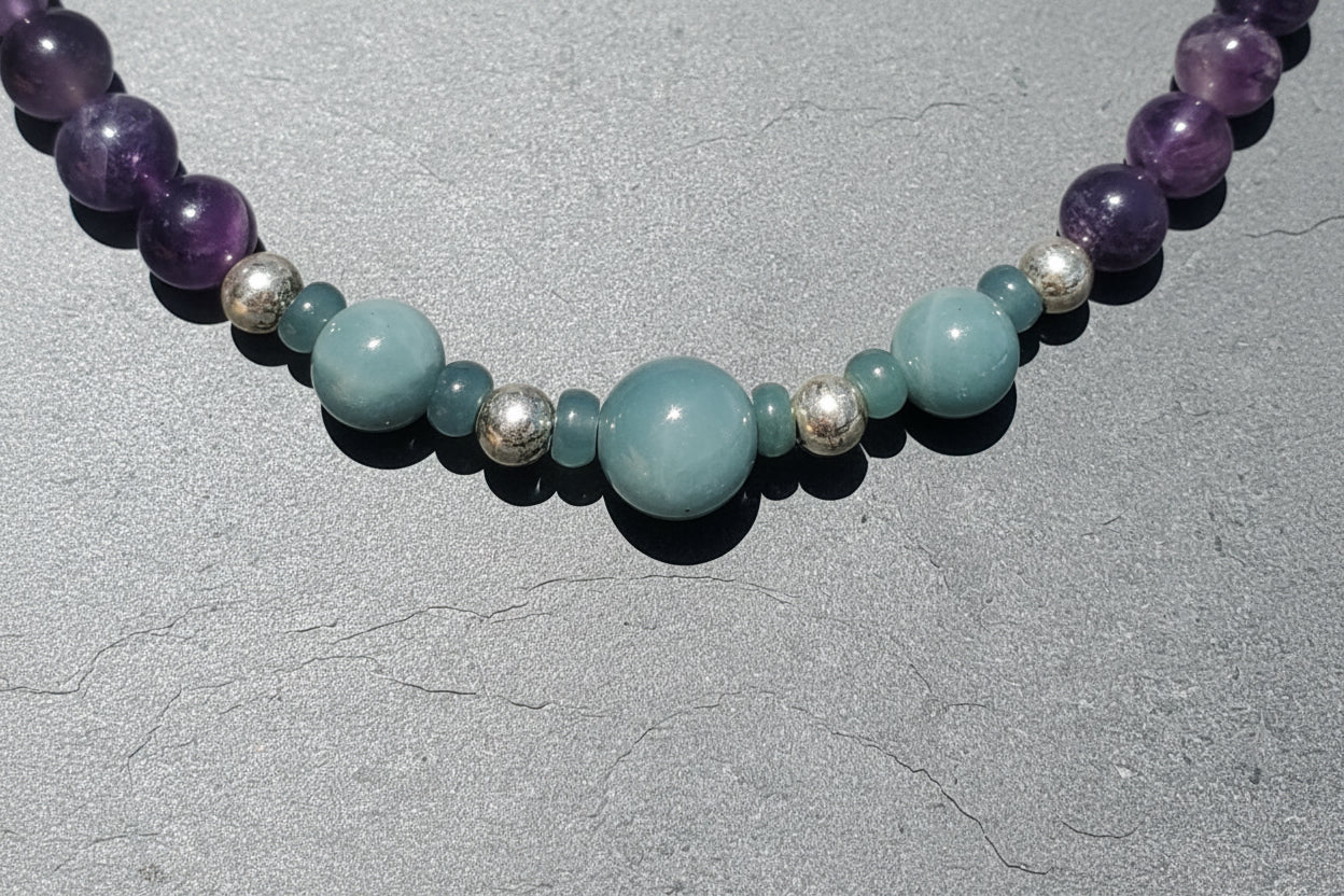 A beaded necklace featuring large light blue-green round beads, smaller matching beads, polished purple amethyst beads, blue jade and metallic silver accent beads, displayed on a textured gray surface.